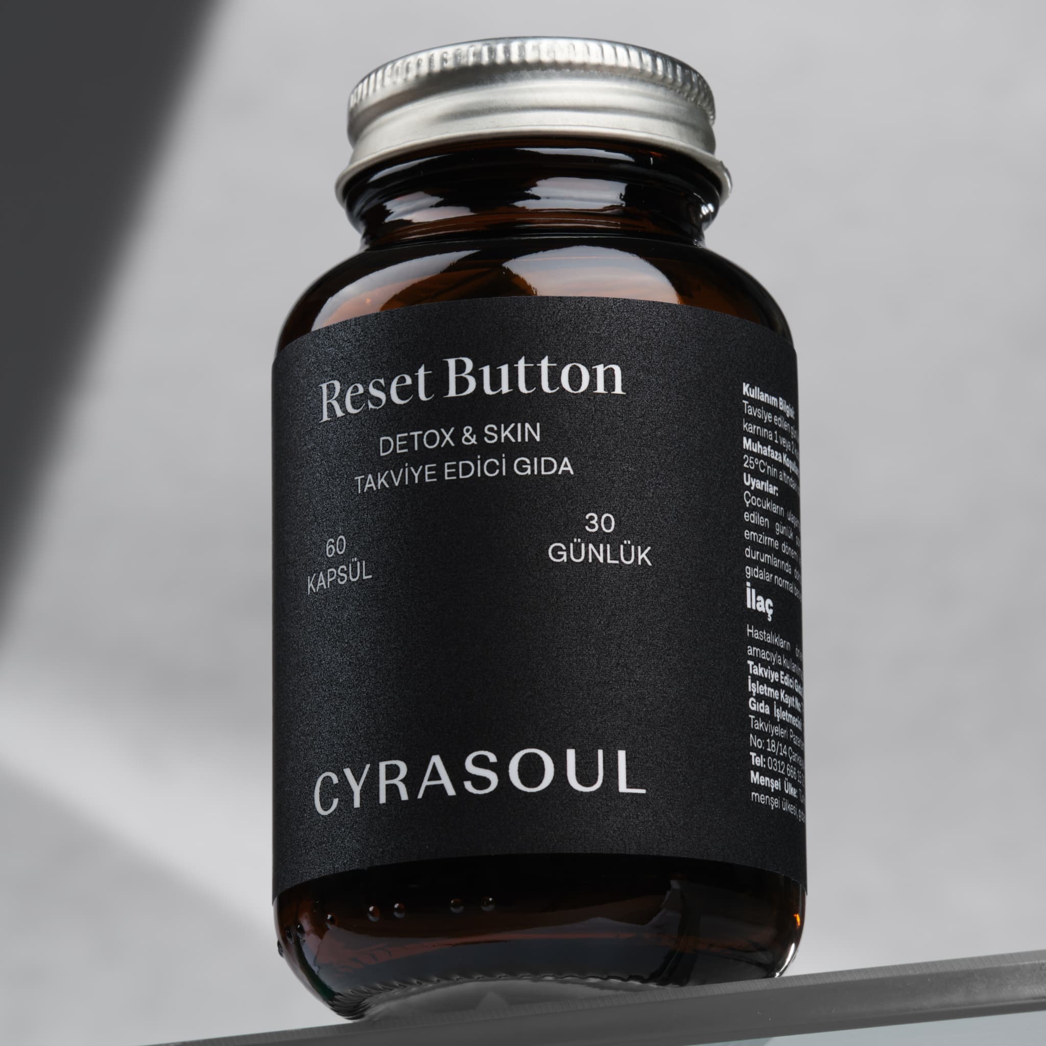 Cyrasoul Reset Button detoks ve cilt takviyesinin gümüş kapaklı amber renkli şişesinin profesyonel stüdyo çekimi. Ürünün premium ve minimalist ambalajı öne çıkıyor.