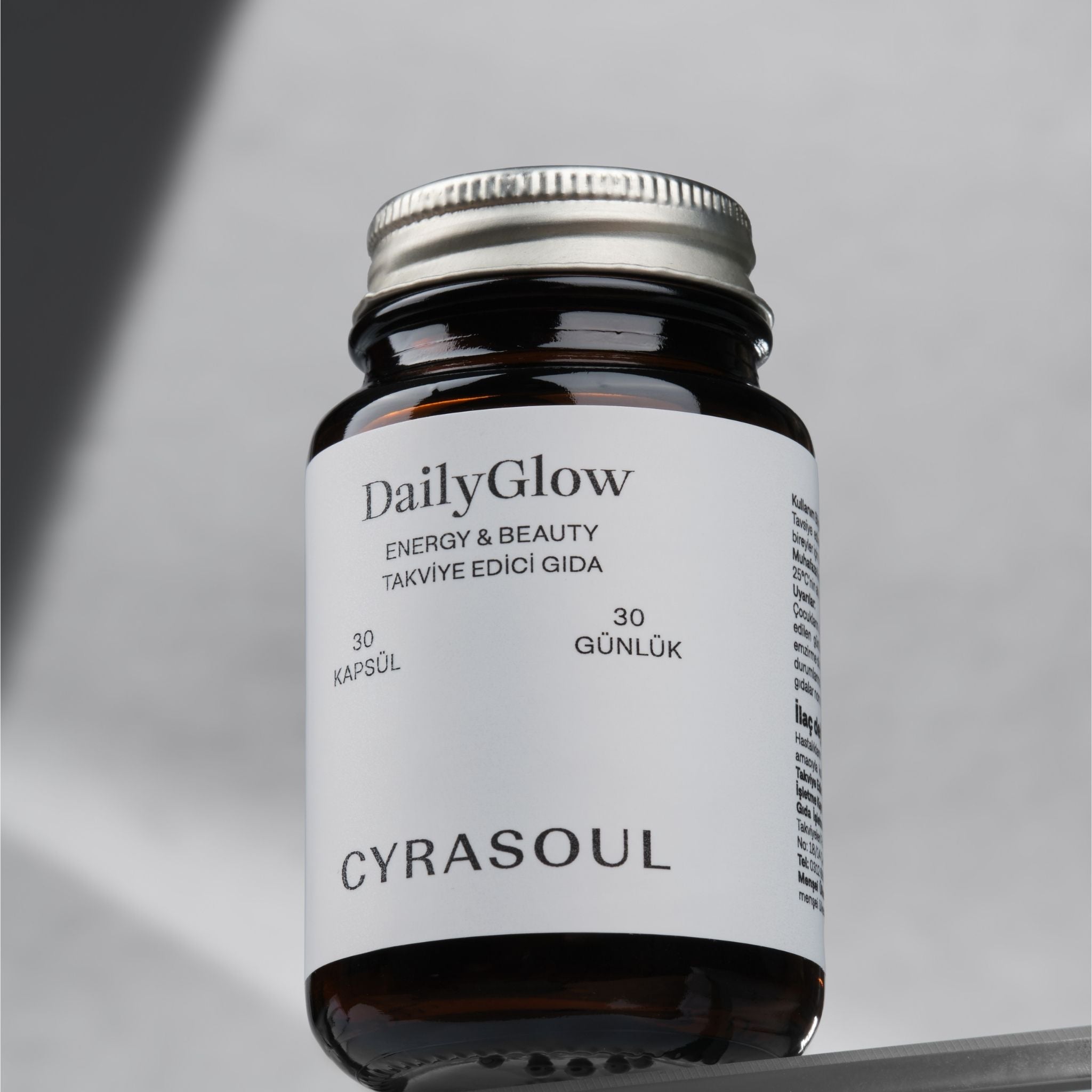 Cyrasoul DailyGlow enerji ve güzellik takviyesinin gümüş kapaklı amber renkli şişesinin stüdyo çekimi. Ürünün modern ve minimalist tasarımı vurgulanıyor.